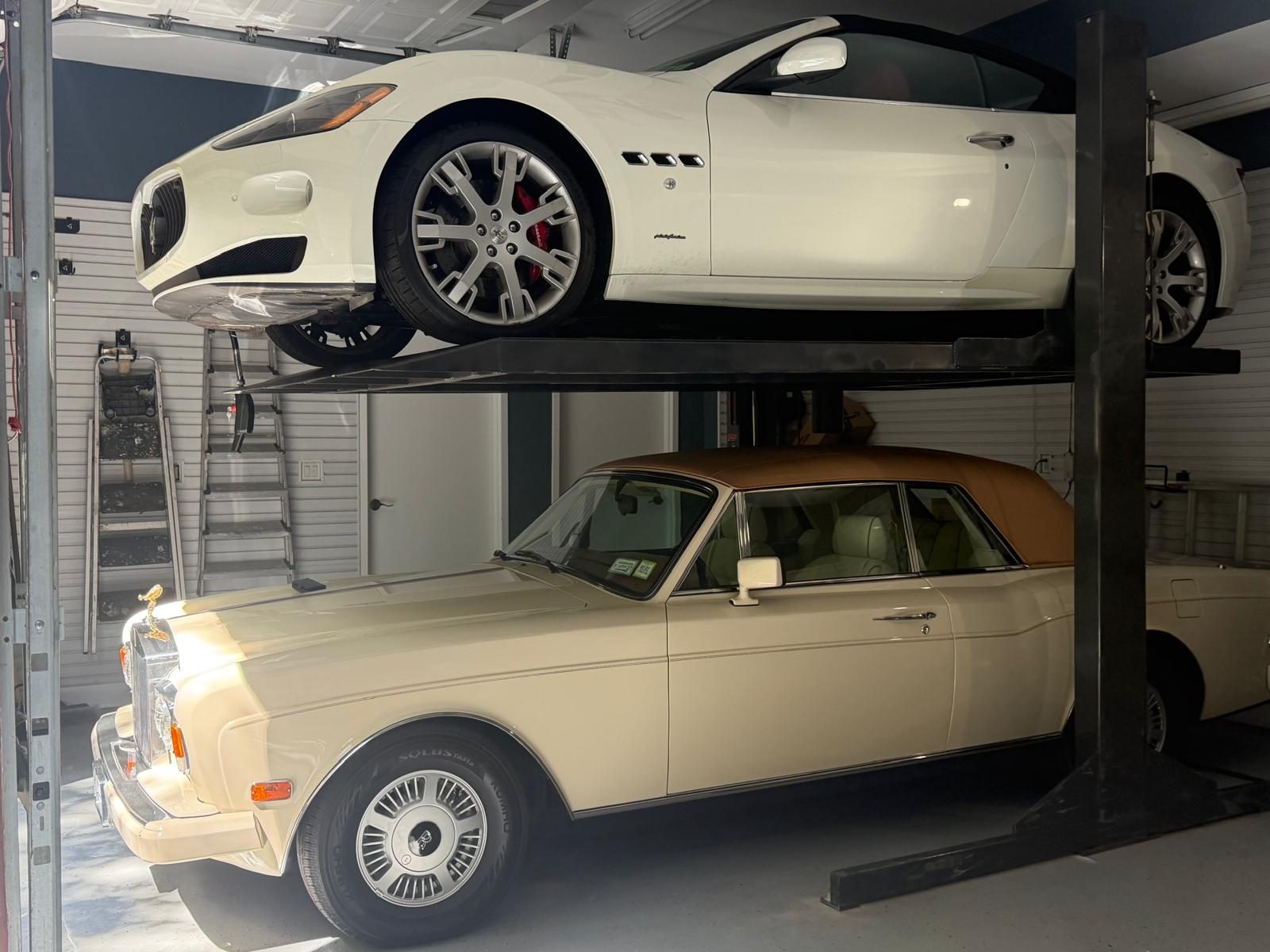 A white coupe parked on the upper platform of a Double Stacker above a classic sedan in a private garage