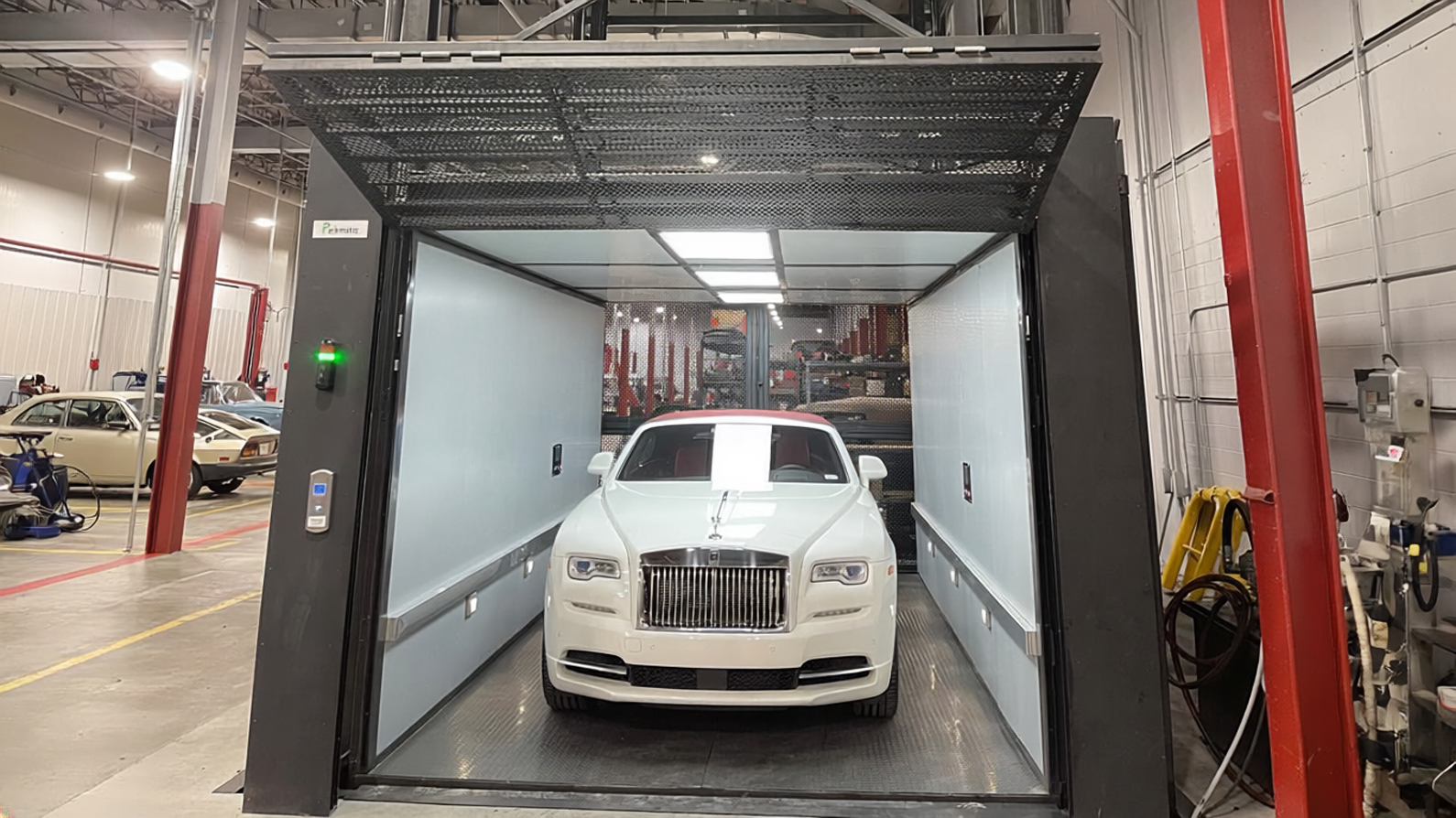 A white sedan parked inside the open cabin of a Parkmatic VRC lift on a workshop floor